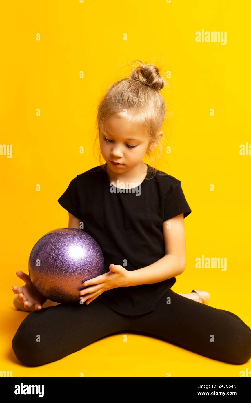 Young beautiful white caucasian girl gymnast doing gymnastic exercise with a sports ball on yellow background Stock Photo
