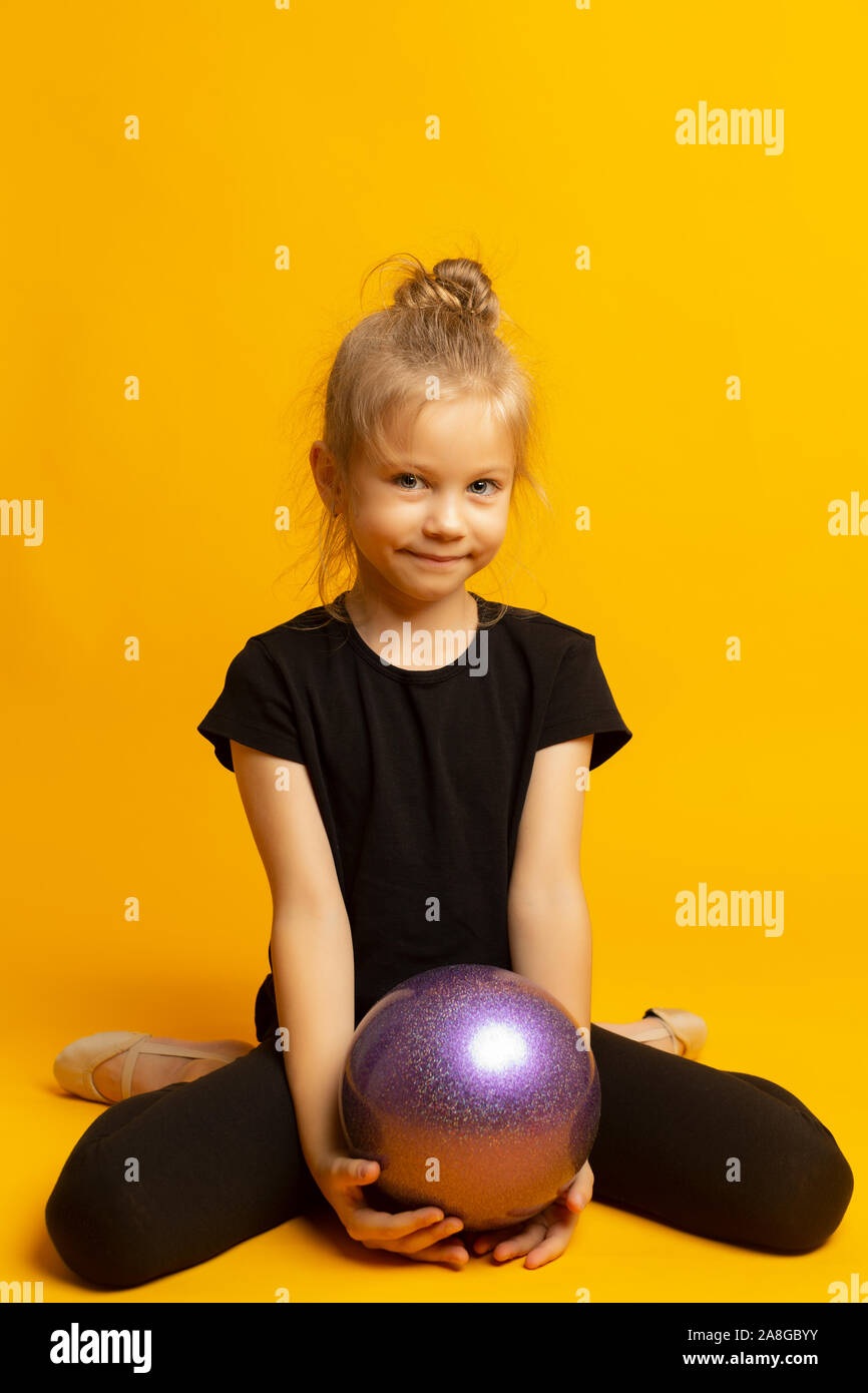 gymnast girl in black trico full height sits on a half page with a sports ball isolated on a yellow background Stock Photo