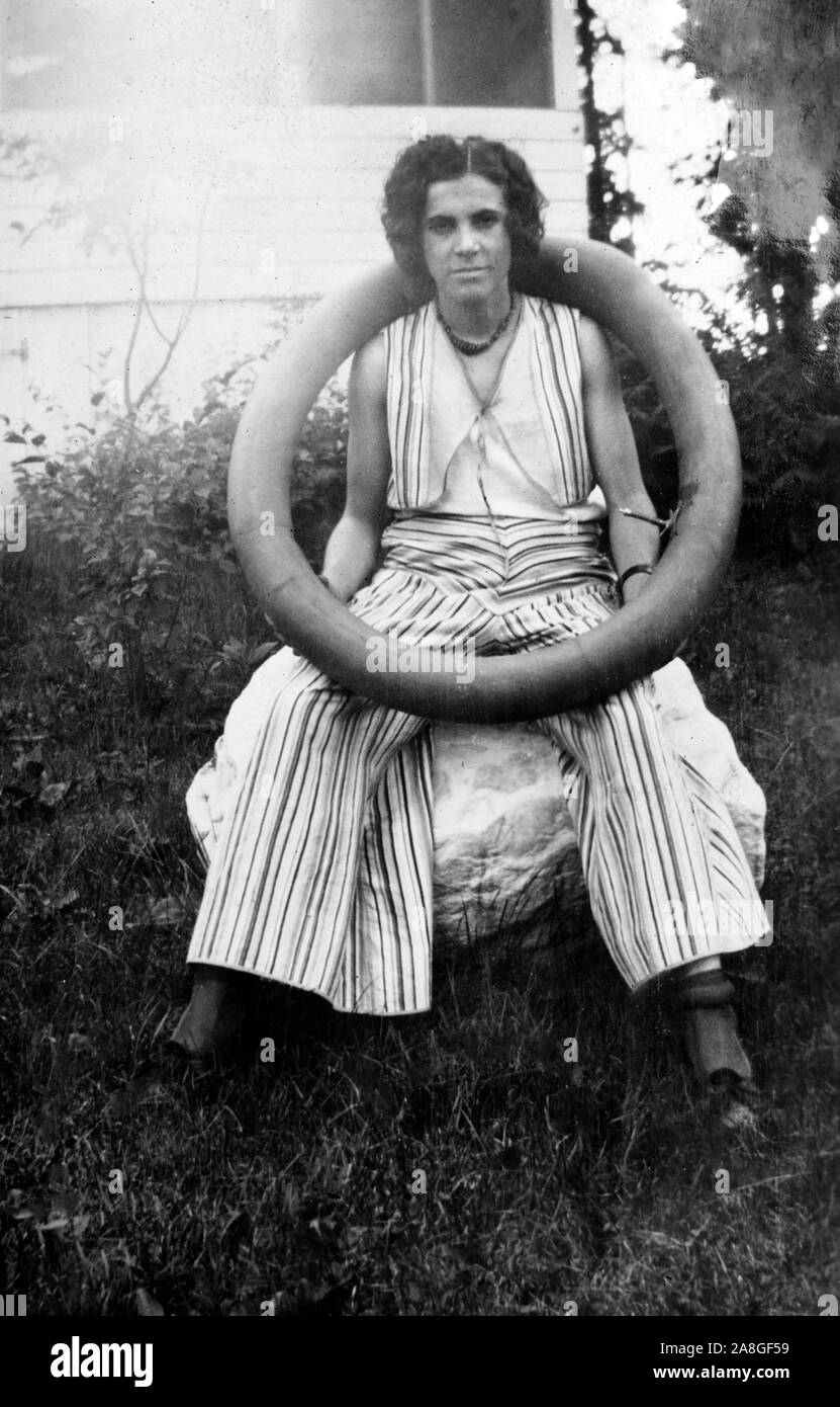 A young woman looks unhappy while sitting on a rock with tire inner tube around her neck, ca. 1920. Stock Photo