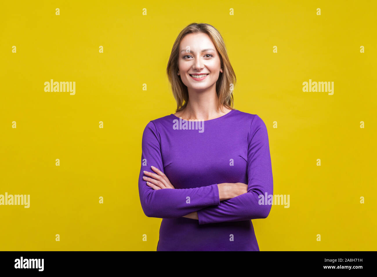 Portrait of happy successful businesswoman in tight purple dress standing with crossed hands, looking at camera with charming and joyous toothy smile. Stock Photo