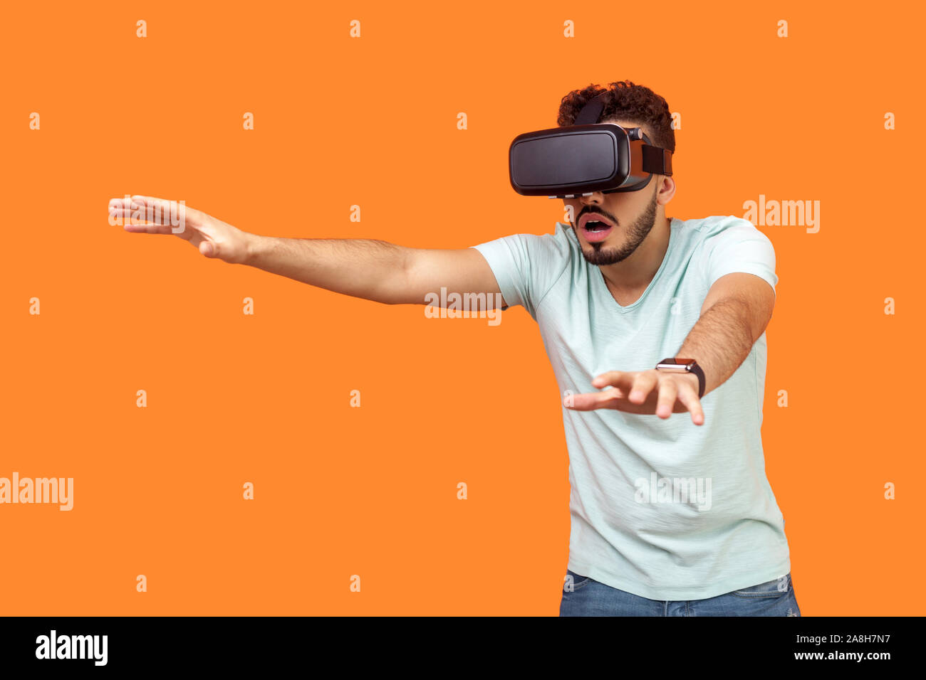 Portrait of scared gamer, brunette man with beard in t-shirt wearing vr glasses, stretching arms forward while playing virtual reality game, frightene Stock Photo