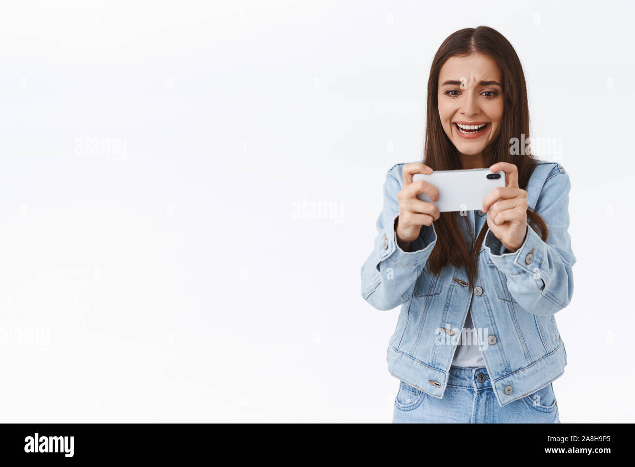 Distressed, intense young attractive woman in denim, holding smartphone horizontally, frowning and grimacing as losing online mobile game battle Stock Photo