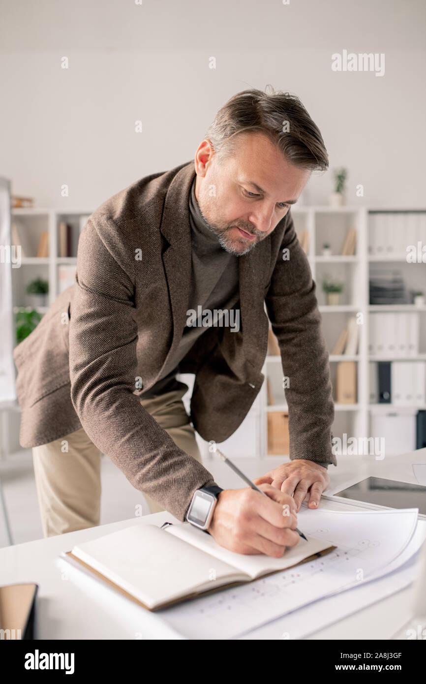 Mature architect bending over open notebook on desk while making working notes Stock Photo