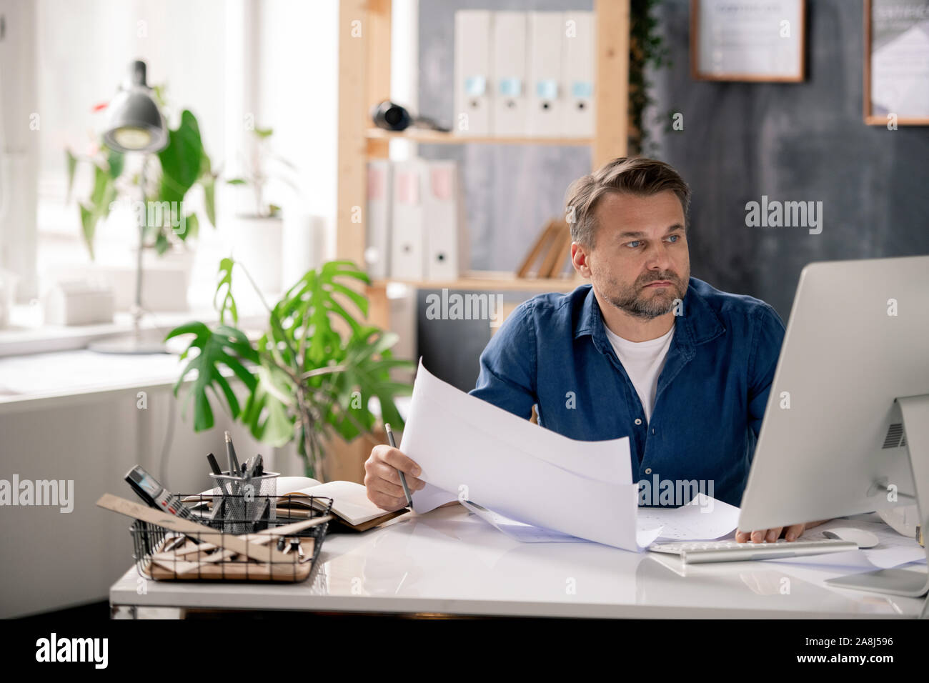 Serious engineer with blueprint looking through data on computer screen Stock Photo