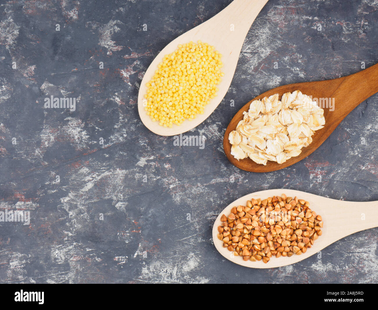 Groats buckwheat, oatmeal, millet in wooden spoons on a dark concrete