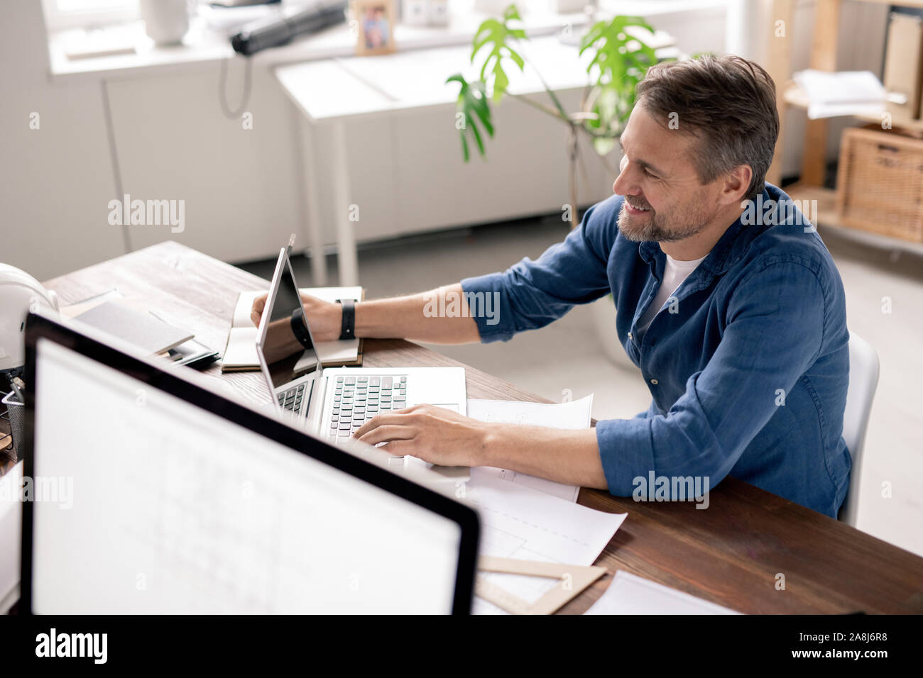 Happy engineer sitting in front of laptop, surfing in the net and making notes Stock Photo