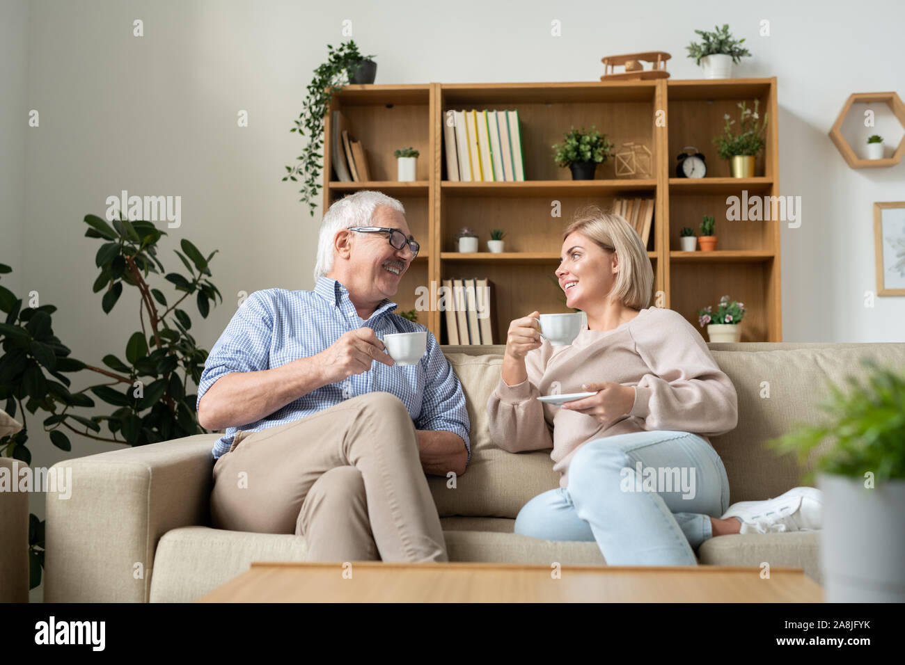 Happy female and her senior father having tea while sitting on couch and talking Stock Photo