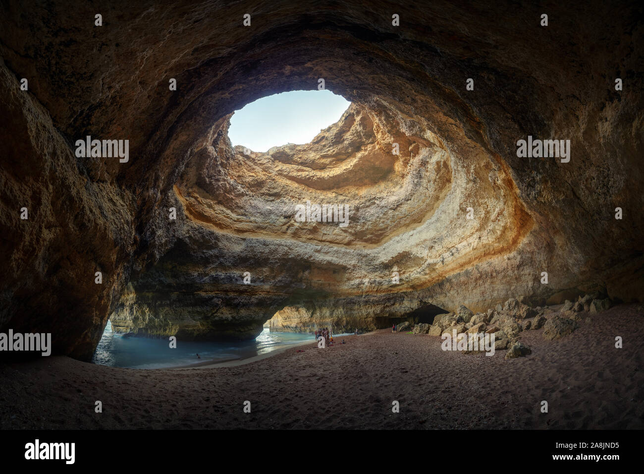 Beach inside a rock cave in Algarve, Portugal. Hole in the ceiling. Blue water and yellow stone. Rocks and sand. People inside the cave. Stock Photo