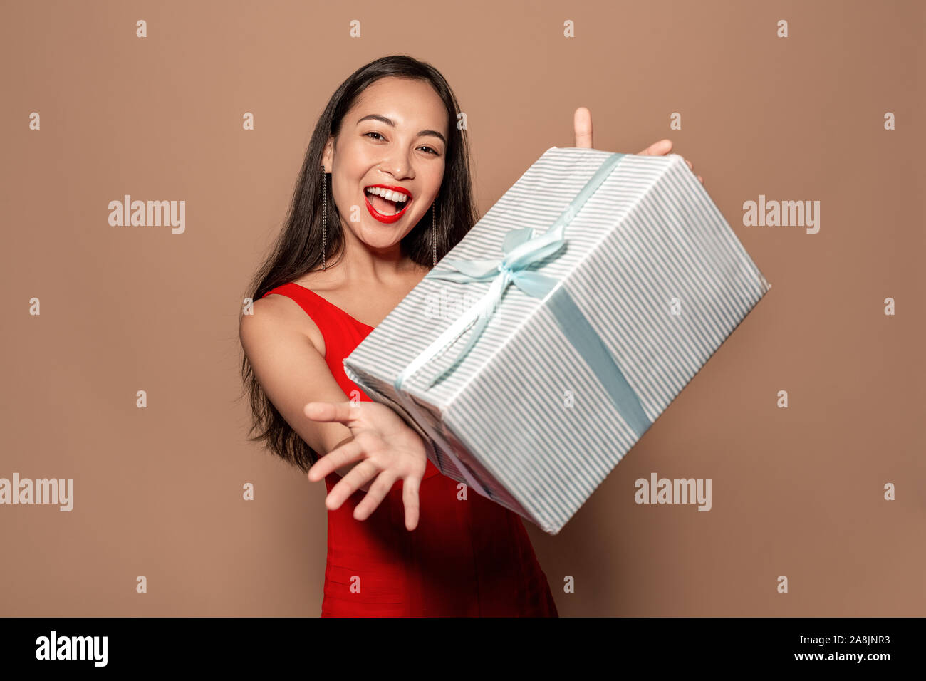 Freestyle. Young girl in dress standing isolated on brown giving present box to camera close-up smiling playful Stock Photo