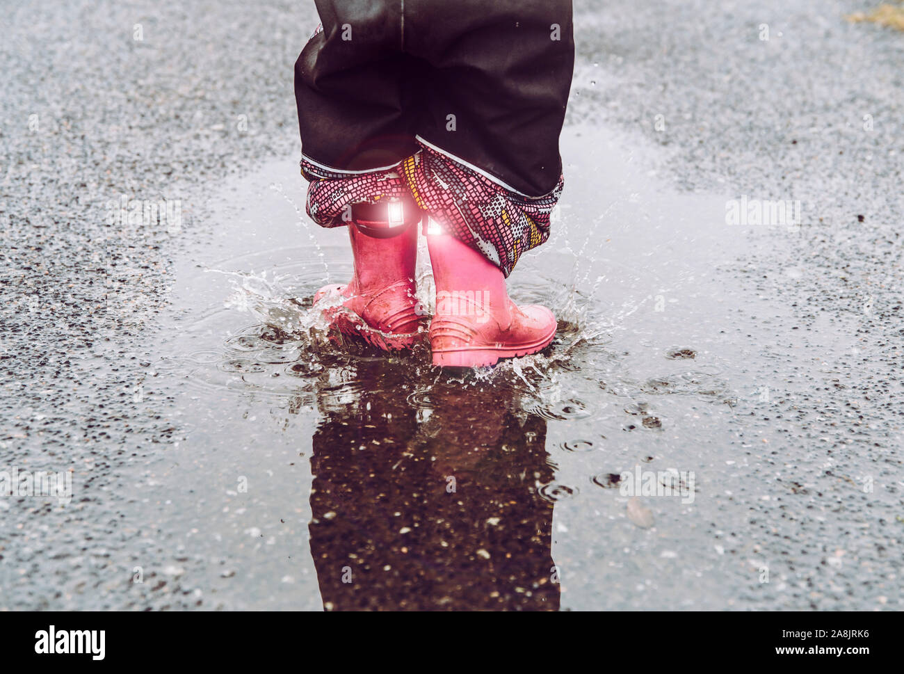 Girl having fun, jumping in water puddle on wet street, wearing rain boots with reflective detail fabric stripes shining. High visibility and safety i Stock Photo