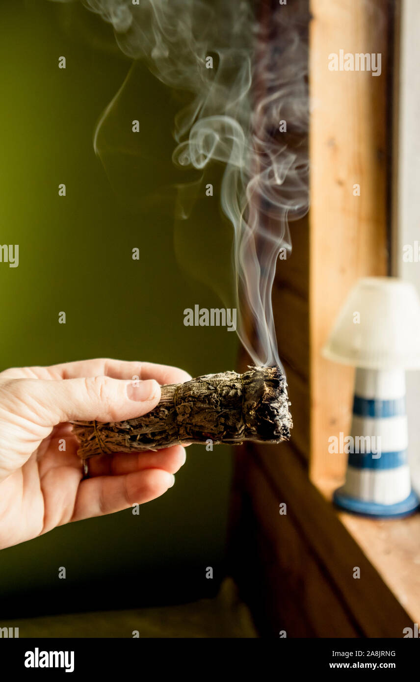 Woman hand holding herb bundle of dried sage smudge stick smoking. It is believed to cleanse negative energy and purify living spaces at home in rooms Stock Photo