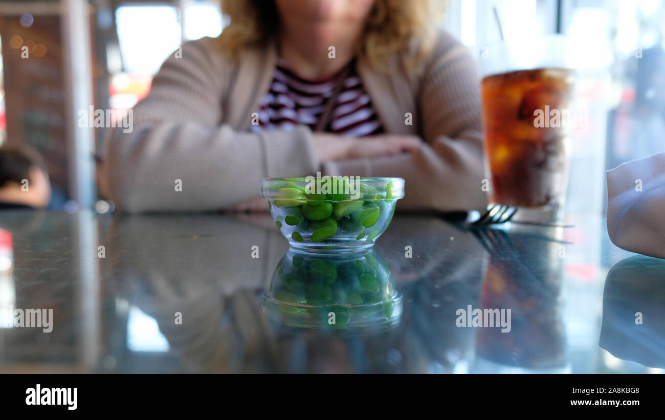 A glass bowl with shelled edamame soy beans on a restaurant table top in front of a woman. Stock Photo