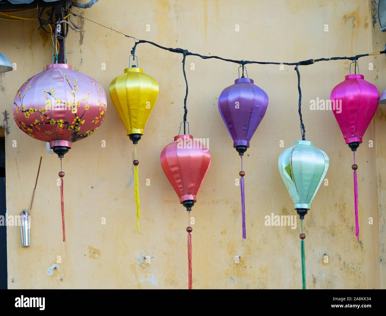 Six satin fabric lanterns in pink, yellow, purple, and mint green hanging against an weathered, gold stucco building. Stock Photo