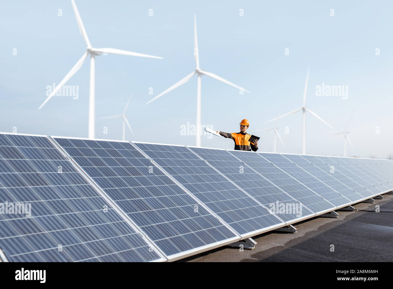 View on the rooftop solar power plant with engineer in protective workwear and windturbines on the background. Concept of alternative energy and its maintenance Stock Photo
