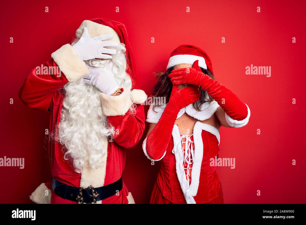 Middle age couple wearing Santa costume and glasses over isolated red background Covering eyes and mouth with hands, surprised and shocked. Hiding emo Stock Photo