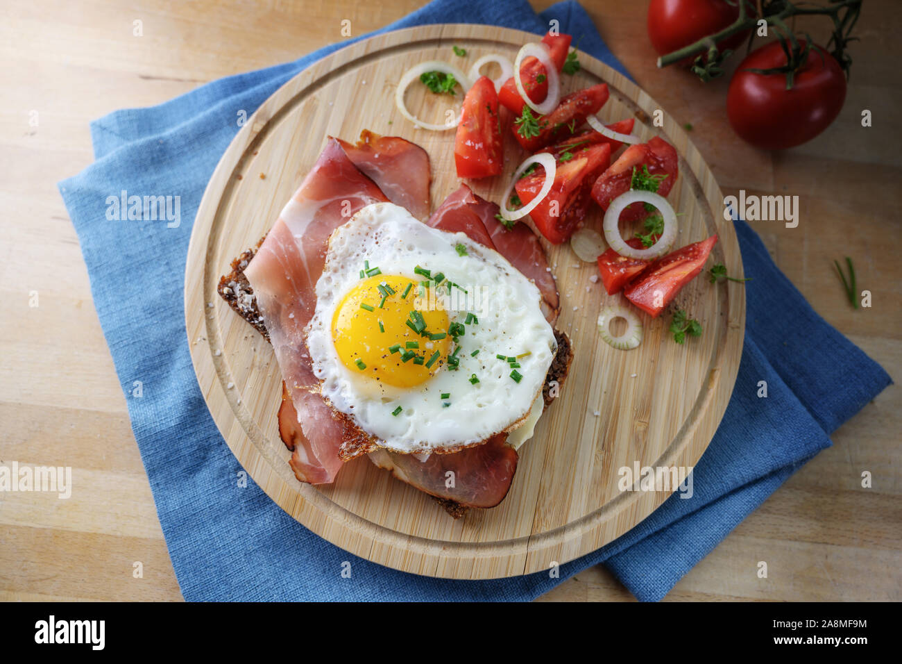 fried egg and ham on a dark whole meal bread, tomatoes, onion rings and herbs garnish, served on a kitchen board and a blue napkin, top view from abov Stock Photo