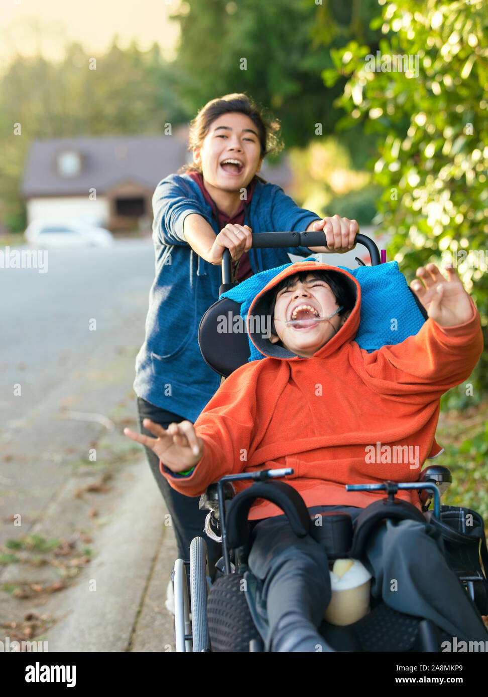 Teen girl running and smiling while pushing disabled little boy in wheelchair outdoors Stock Photo