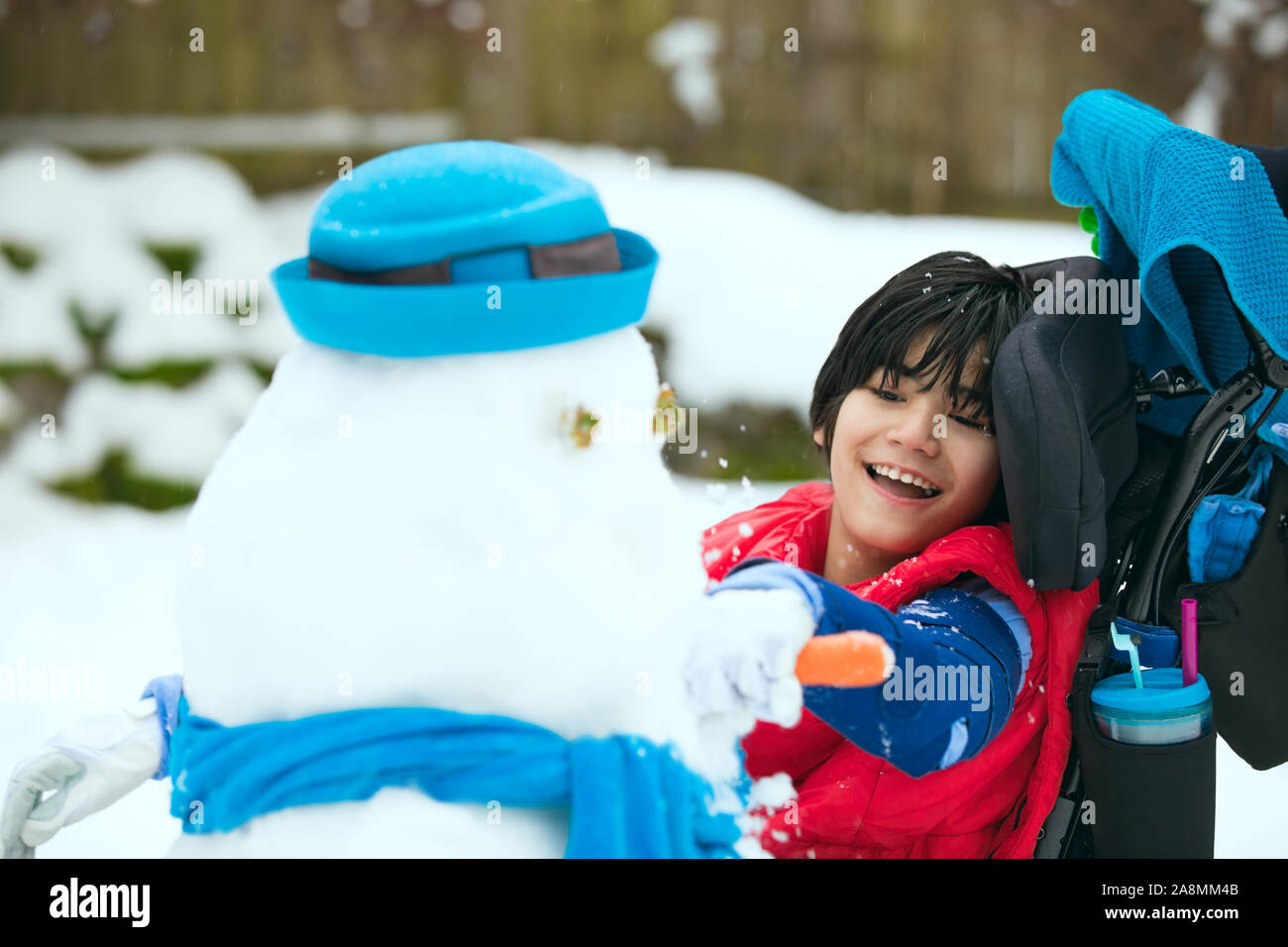 Smiling, happy little boy in wheelchair outdoors in winter having fun building a snowman Stock Photo