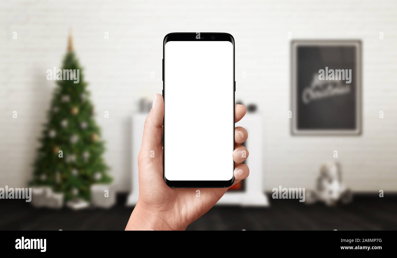 Phone in hand mockup. Christmas scene with decorated Christmas tree, fireplace and poster in background. Stock Photo