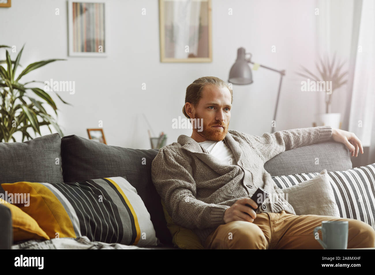 Portrait of bored bearded man watching TV at home and holding remote control while relaxing on comfortable couch, copy space Stock Photo