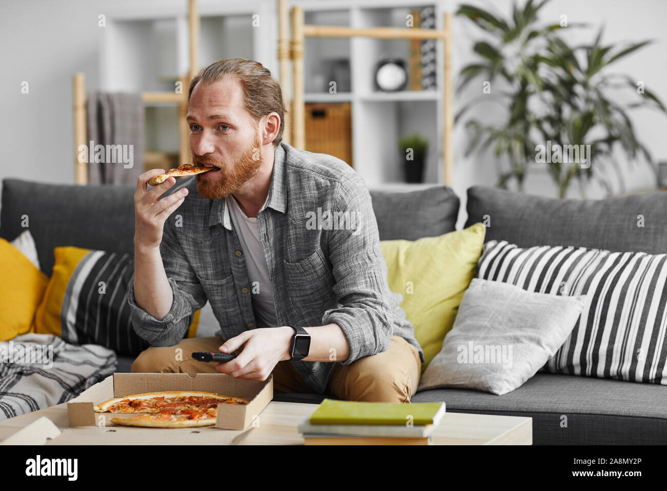 Portrait of middle-aged bearded man watching TV at home and eating pizza during lazy weekend, copy space Stock Photo