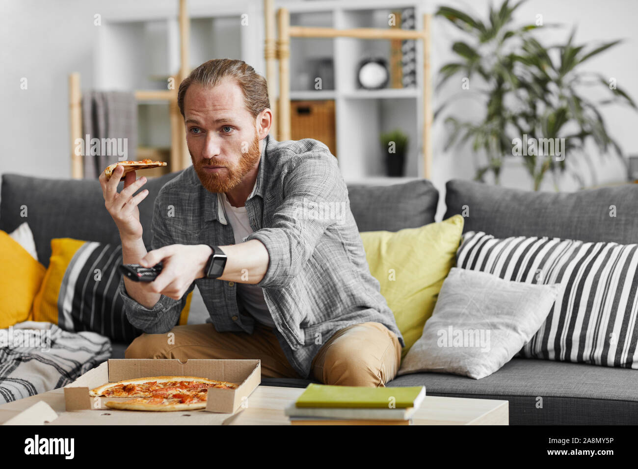 Portrait of middle-aged man watching TV at home and eating pizza during lazy weekend, copy space Stock Photo