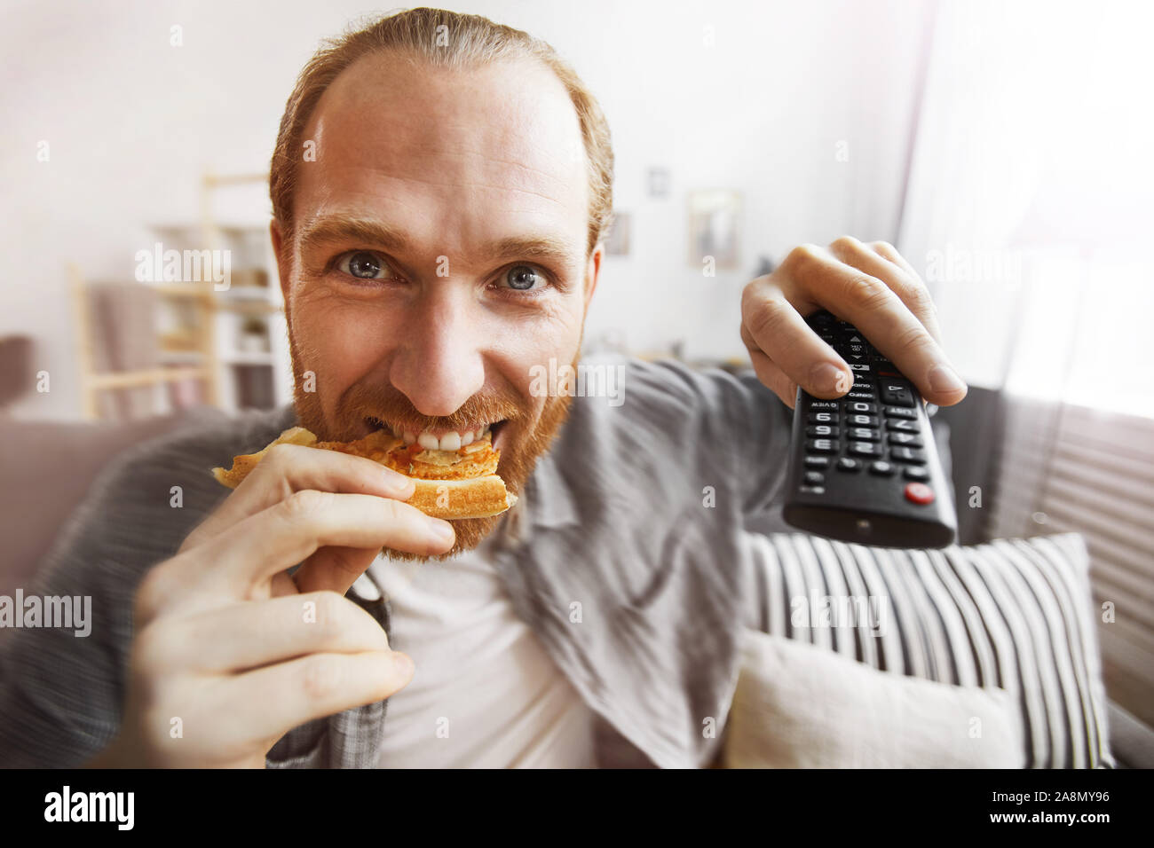 Fish eye portrait of smiling bearded man watching TV at home and eating pizza while binge watching favorite series, copy space Stock Photo