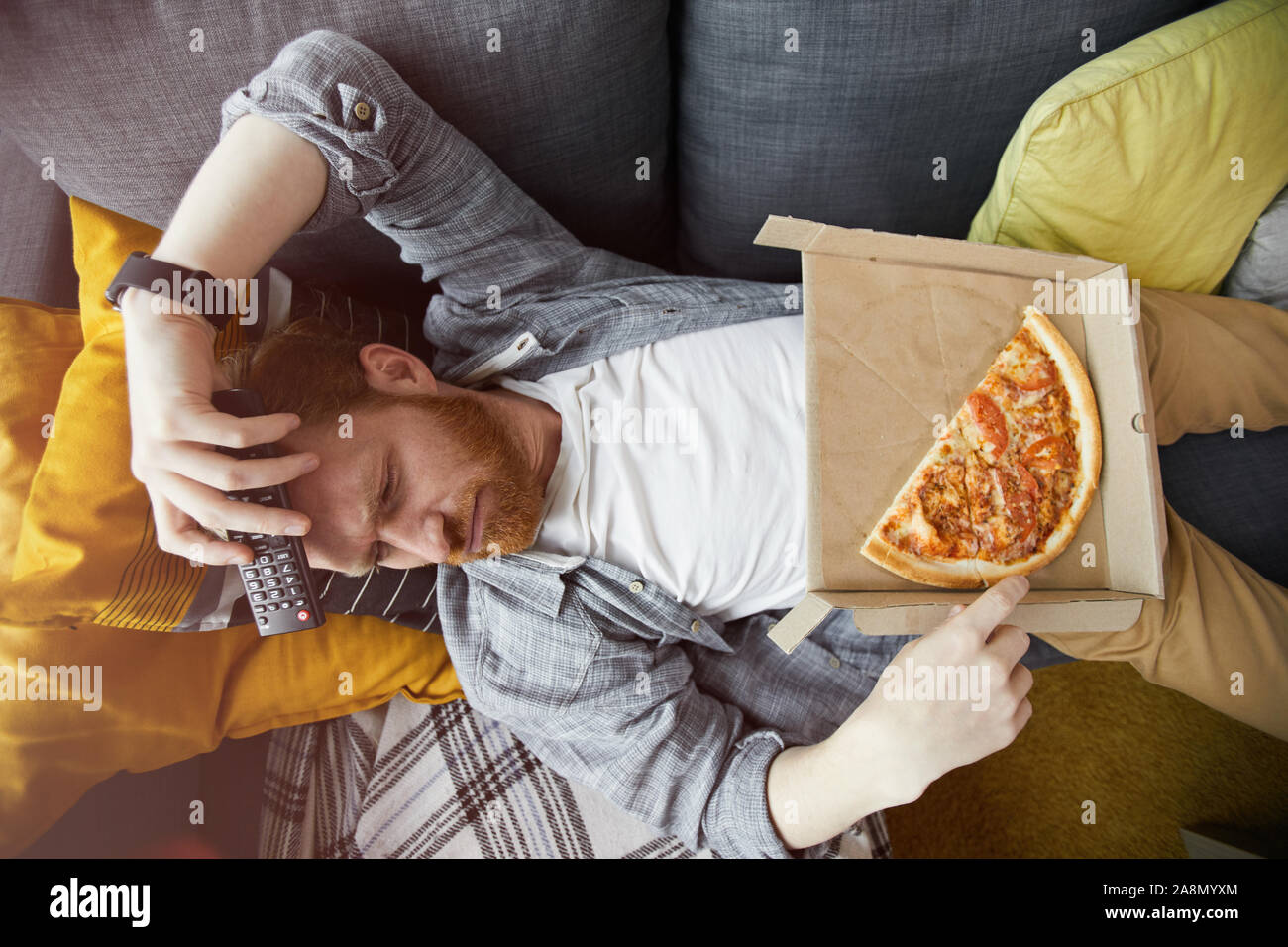 Above view portrait of bearded man lying on couch and eating pizza while watching TV at home, copy space Stock Photo