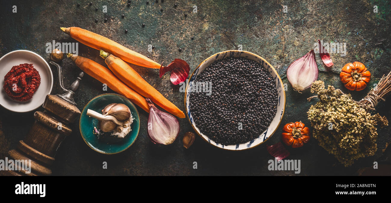 Black lentils with cooking ingredients, vegetables, herbs and spices for tasty vegan meals on dark background. Top view. Healthy vegetarian eating con Stock Photo