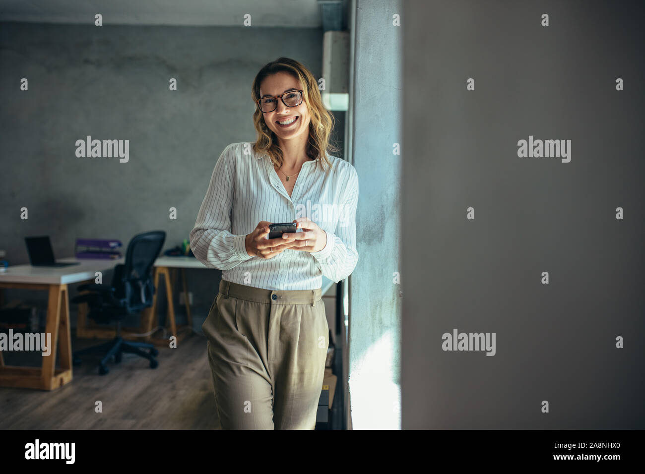 Cheerful woman standing in office with her phone. Businesswoman with cell phone in hand looking at camera and smiling. Stock Photo