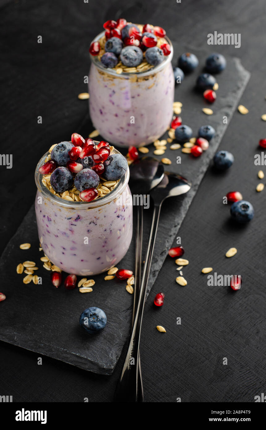 Healthy breakfast concept. Jars of homemade yogurt with blueberries, pomegranate seeds and oats on black background. Vertical. Stock Photo