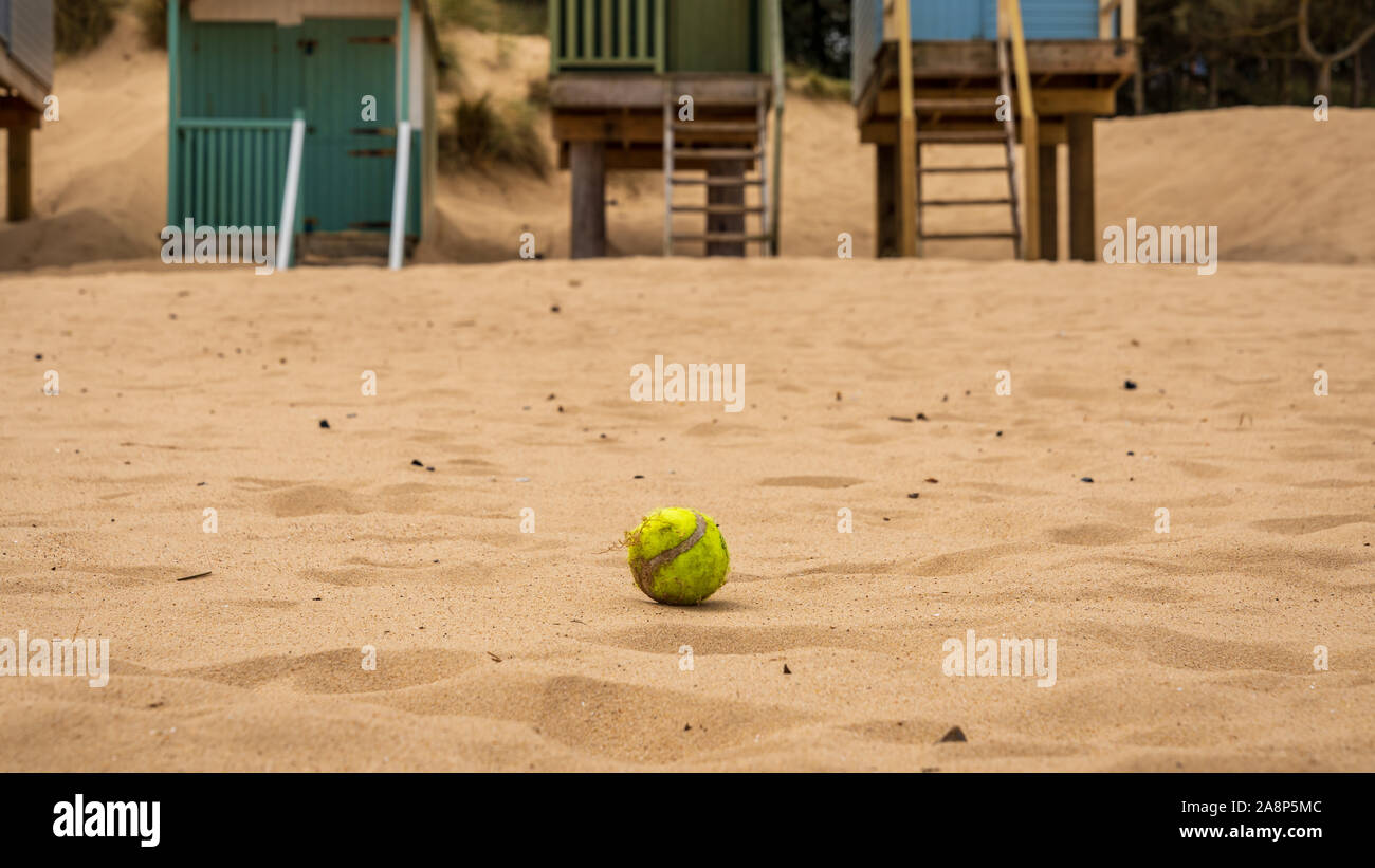 A tennis ball in the sand of the beach, with Beach Huts in the blurry