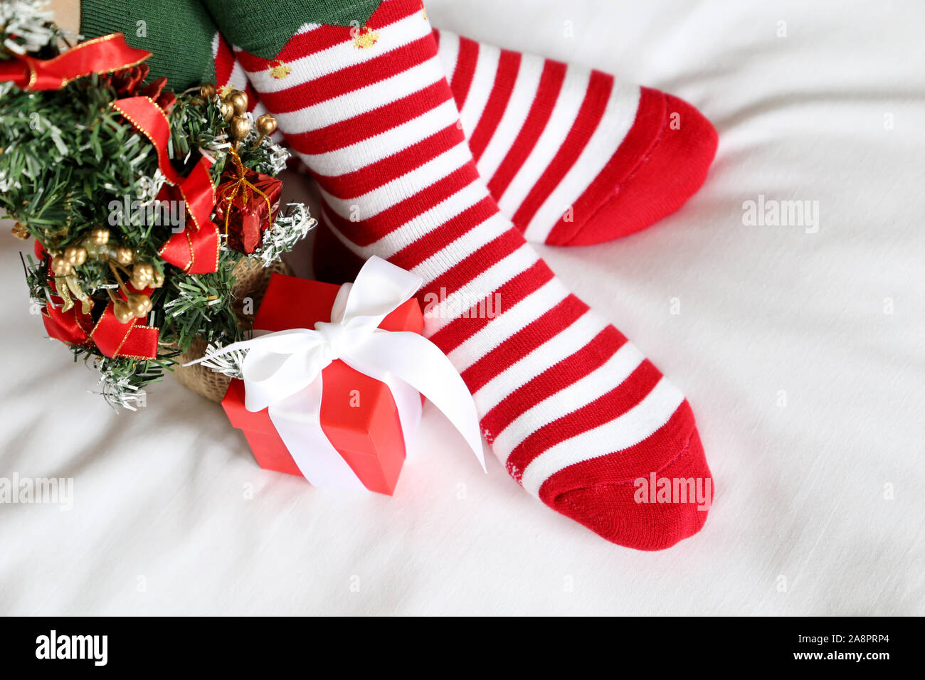 Christmas celebration at home, crossed female feet in red and white striped socks and New Year tree on a bed. Woman in Santa clothes and festive decor Stock Photo