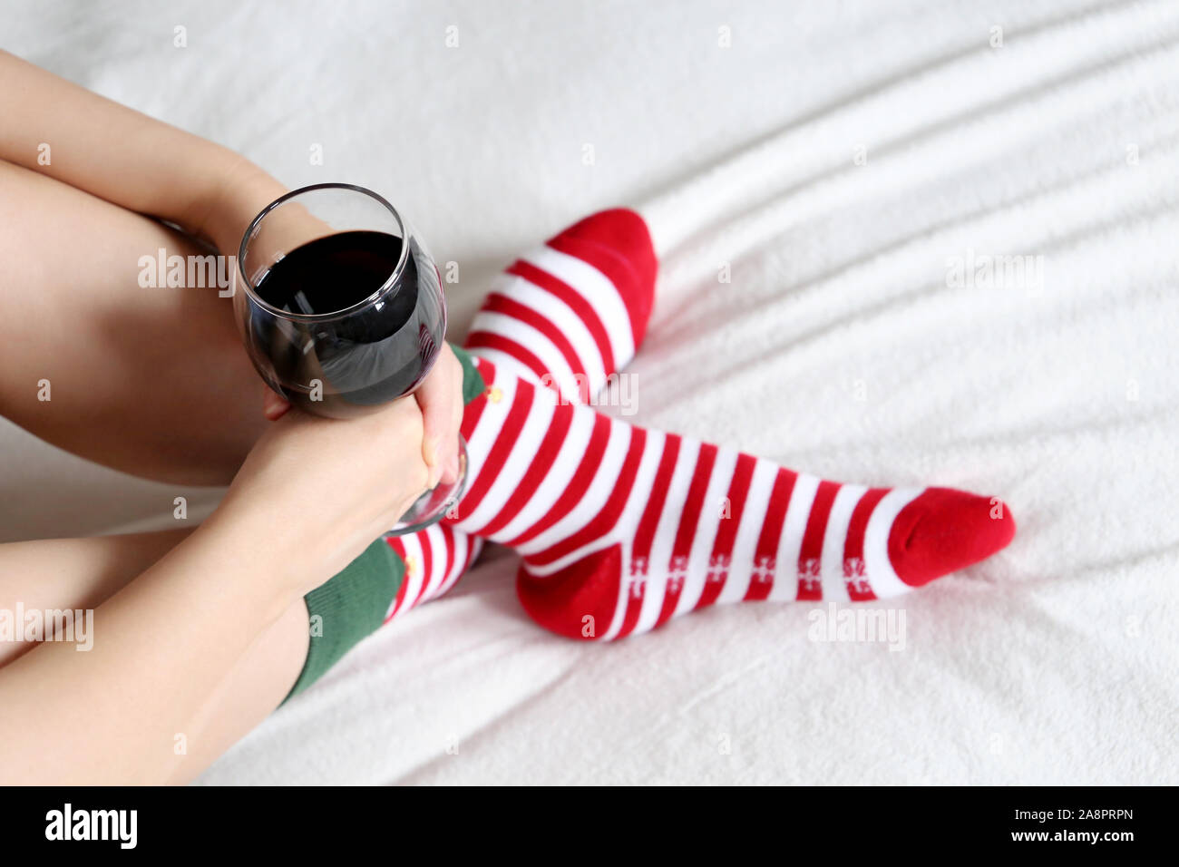 Woman drinking red wine sitting on a bed cross-legged in Christmas socks. Concept of New Year celebration, home relax and cozy atmosphere Stock Photo