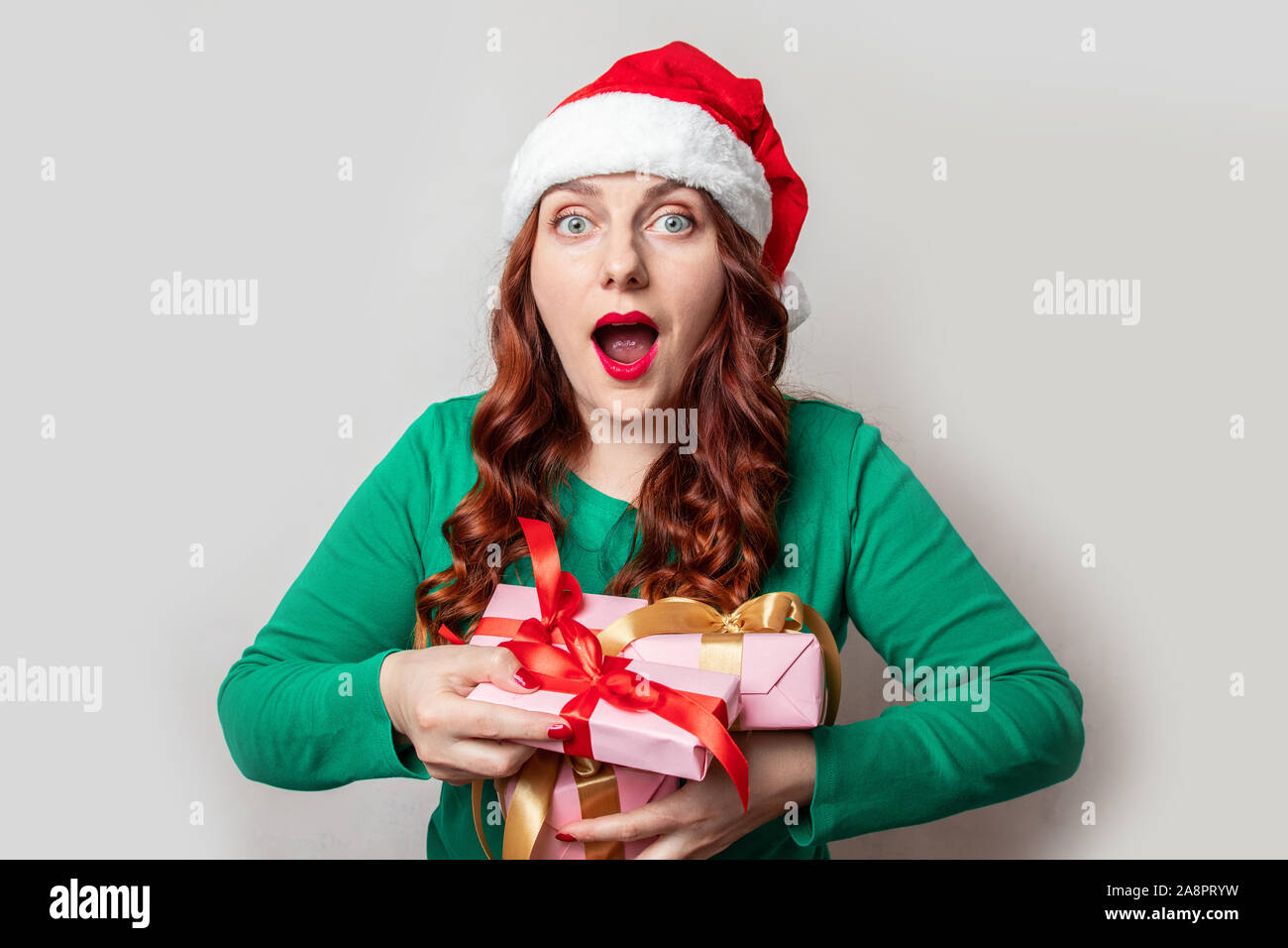 Young beautiful woman wearing red santa hat with funny expression and holds gift boxes on gray background for holiday shopping on boxing day Stock Photo