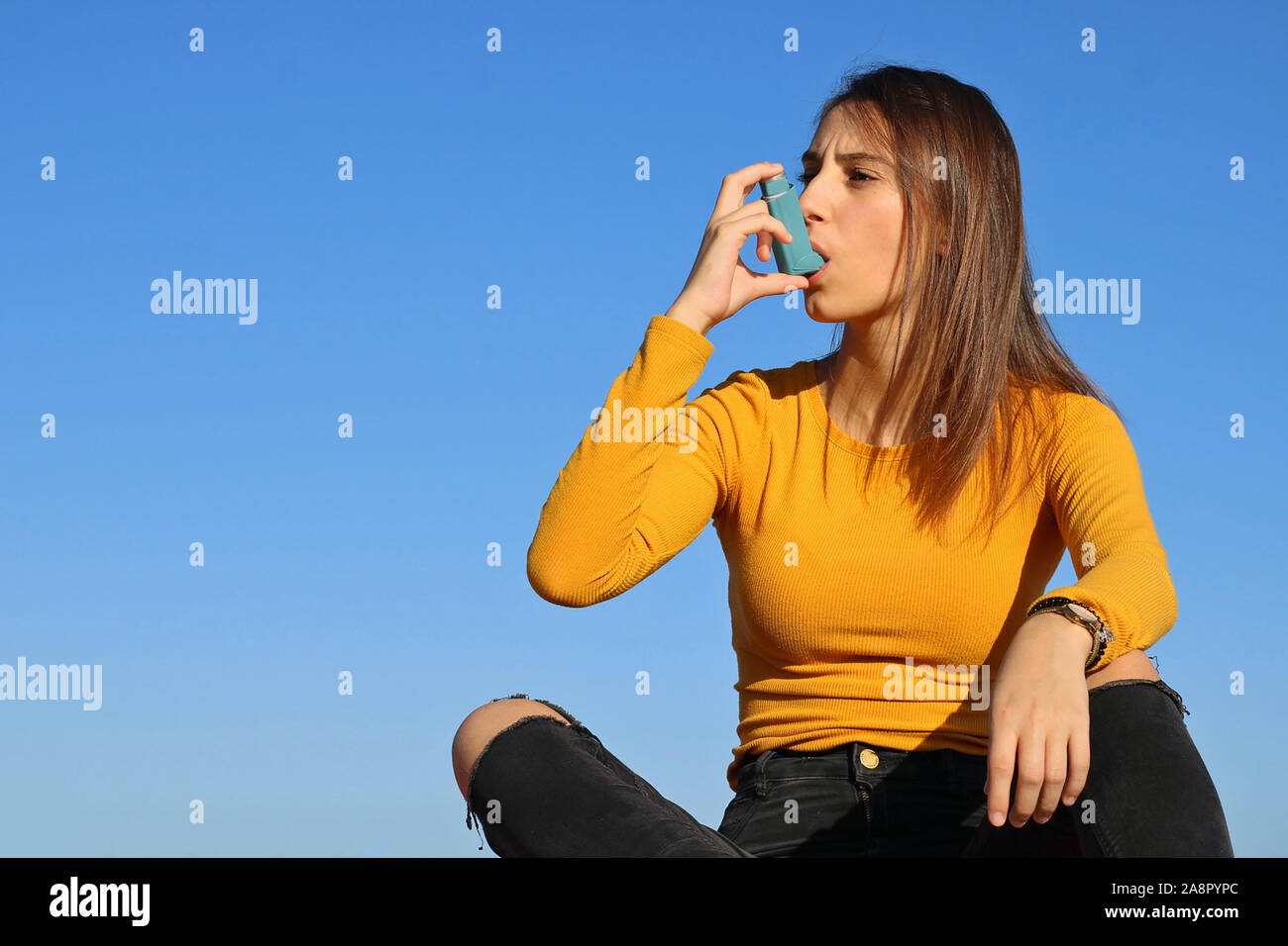 Beautiful young girl dressed in yellow taking the asthma inhaler on a blue background. Pharmaceutical product to prevent an asthma attack. Stock Photo