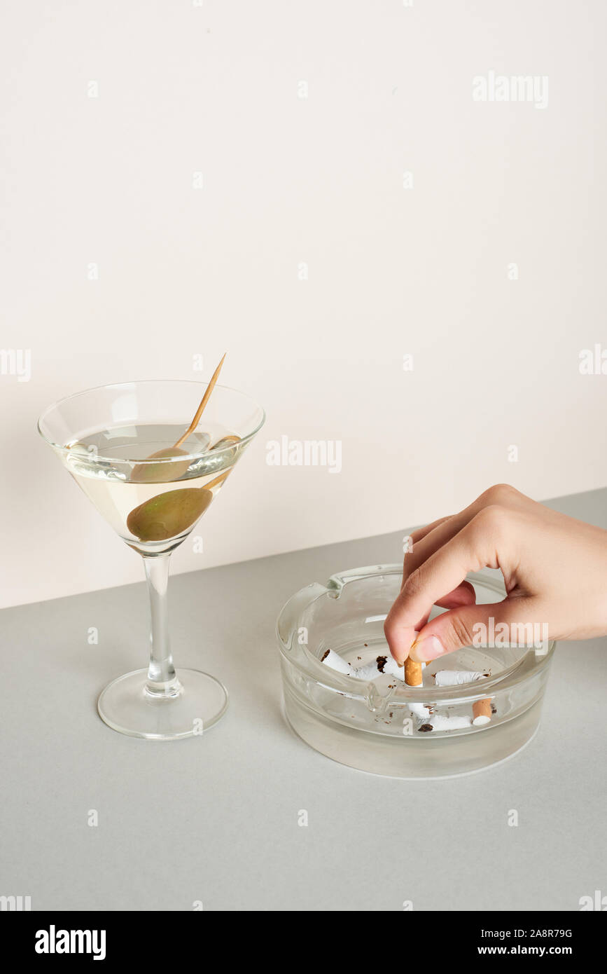 Cropped view of woman putting cigarette to ashtray with martini cocktail on  grey surface Stock Photo