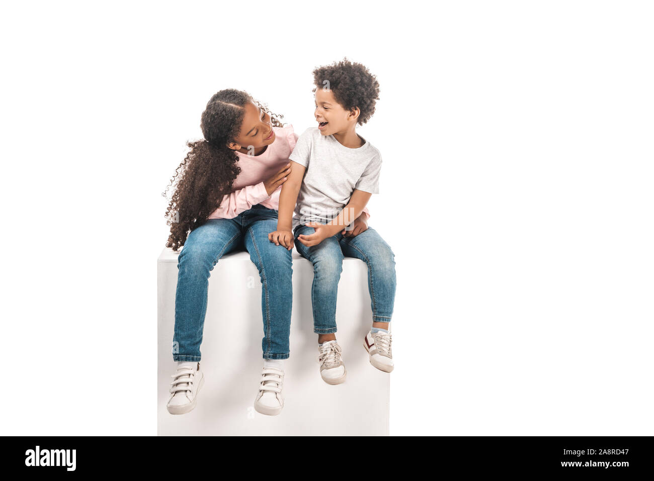 cute african american sister looking at adorable brother while sitting on white cube together isolated on white Stock Photo