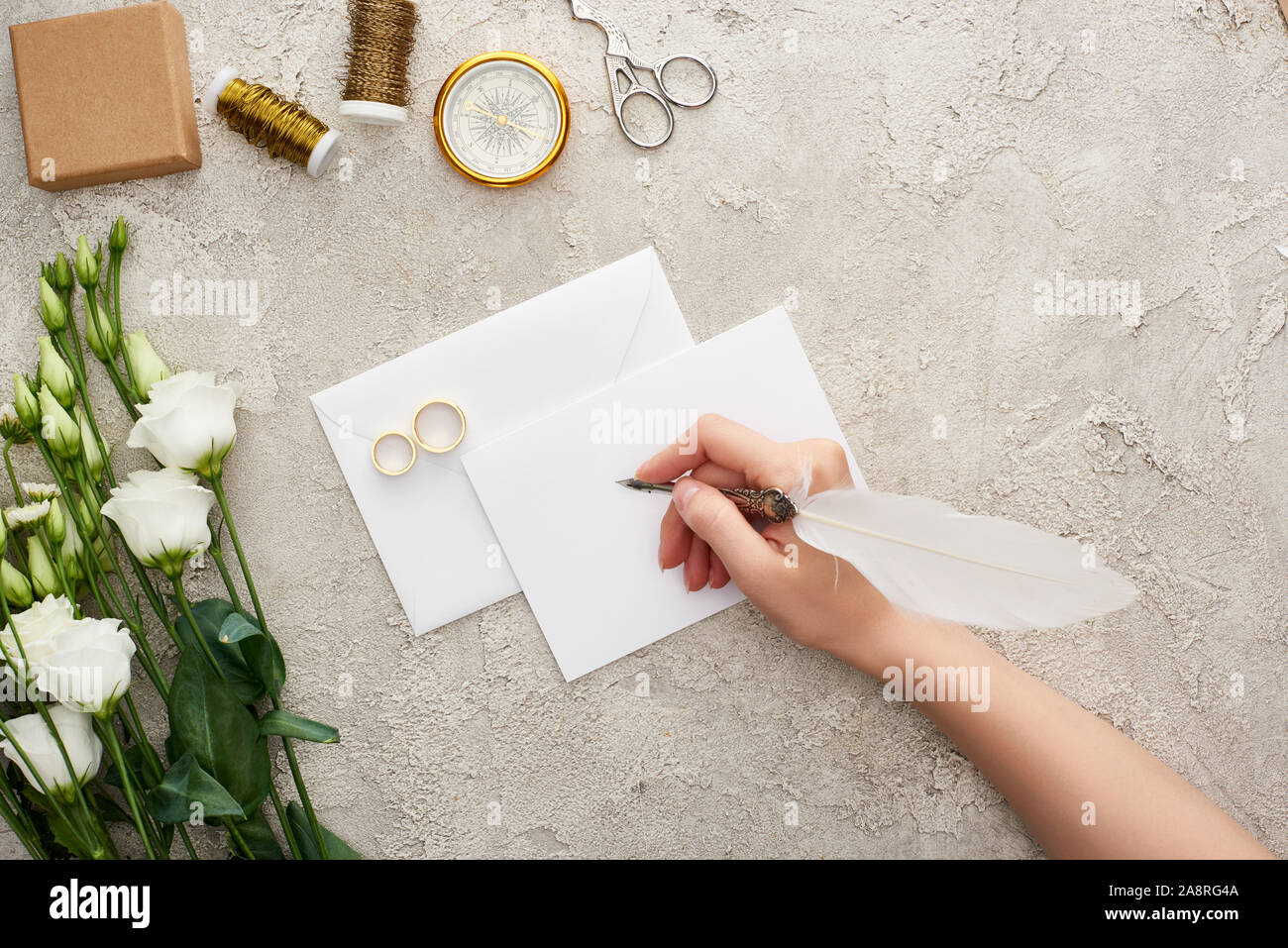cropped view of woman writing on empty card near wedding rings, compass, scissors, bobbins and eustoma flowers on textured surface Stock Photo