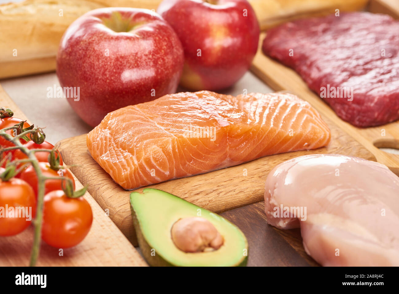 Fresh raw fish, poultry, meat on wooden cutting boards near apples, branch of cherry tomatoes and half of avocado Stock Photo