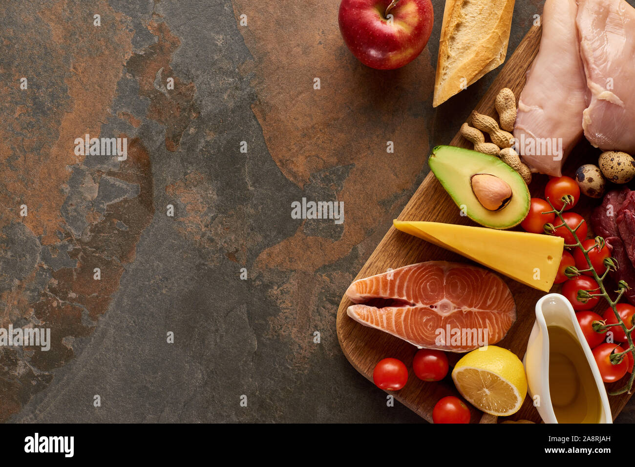 Top view of wooden cutting board with raw fish, poultry, cheese, fruits, vegetables, olive oil, baguette and peanuts on dark brown marble surface with Stock Photo