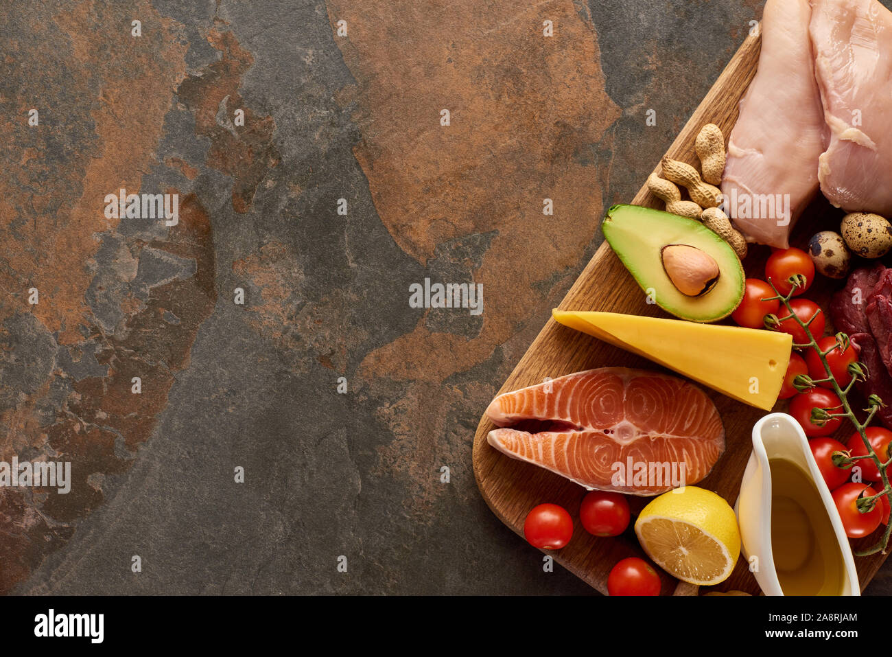 Top view of wooden cutting board with raw fish, poultry, cheese, vegetables, olive oil and peanuts on dark brown marble surface with copy space Stock Photo