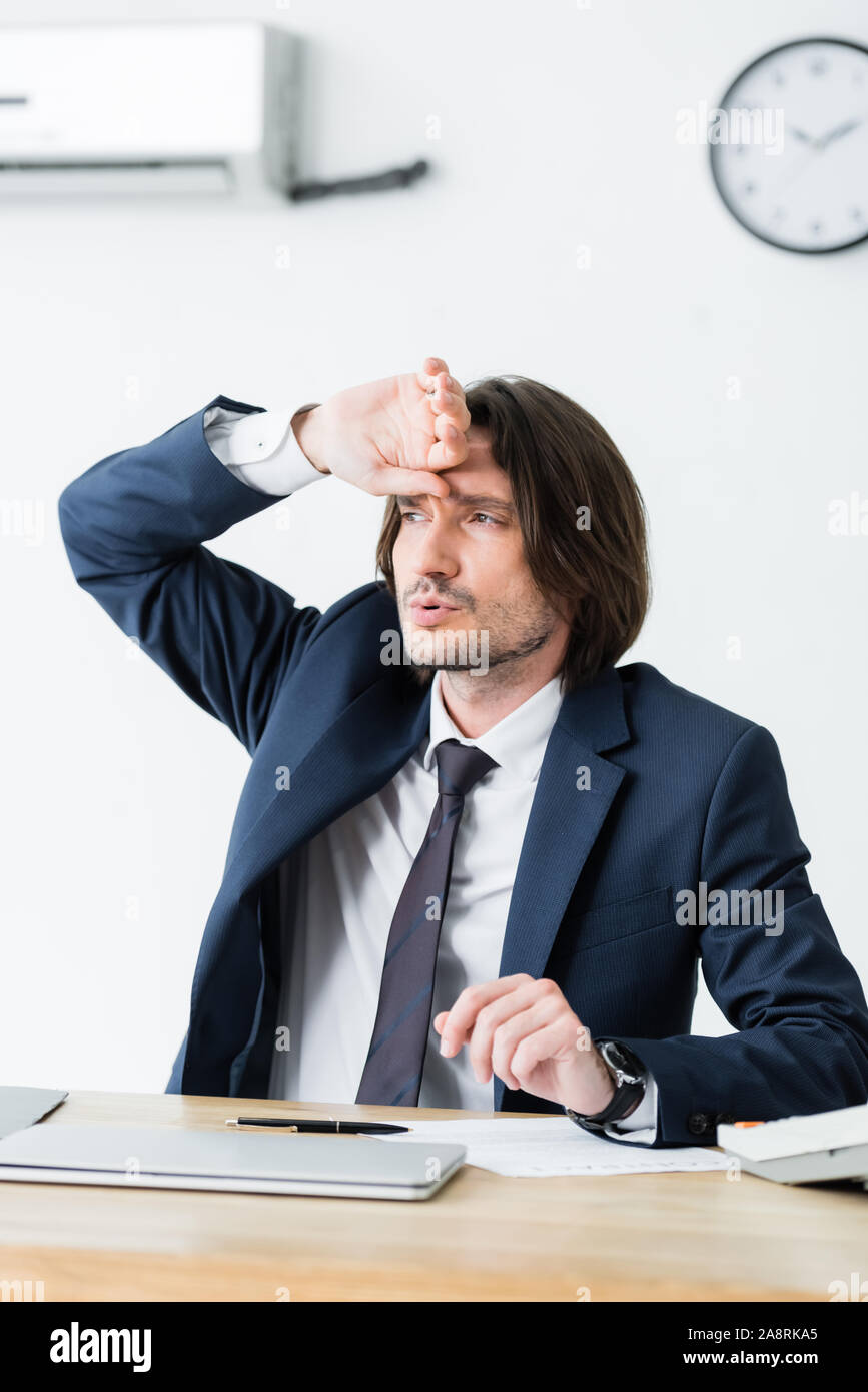 sweaty businessman sitting in office, holding hand near face, looking ...