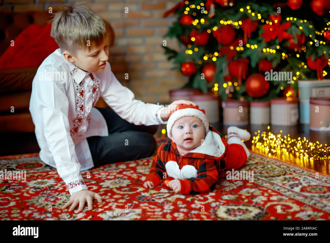 Brother with little sister dressed as Santa Claus. at the New Year tree. Family celebrates Christmas. 6 months old little boy learns to crawl on backg Stock Photo