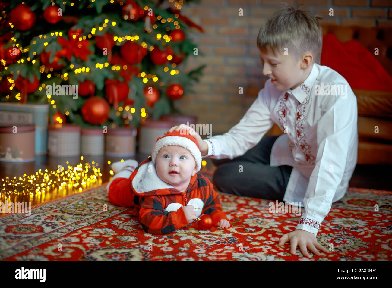Brother with little sister dressed as Santa Claus. at the New Year tree. Family celebrates Christmas. 6 months old little boy learns to crawl on backg Stock Photo