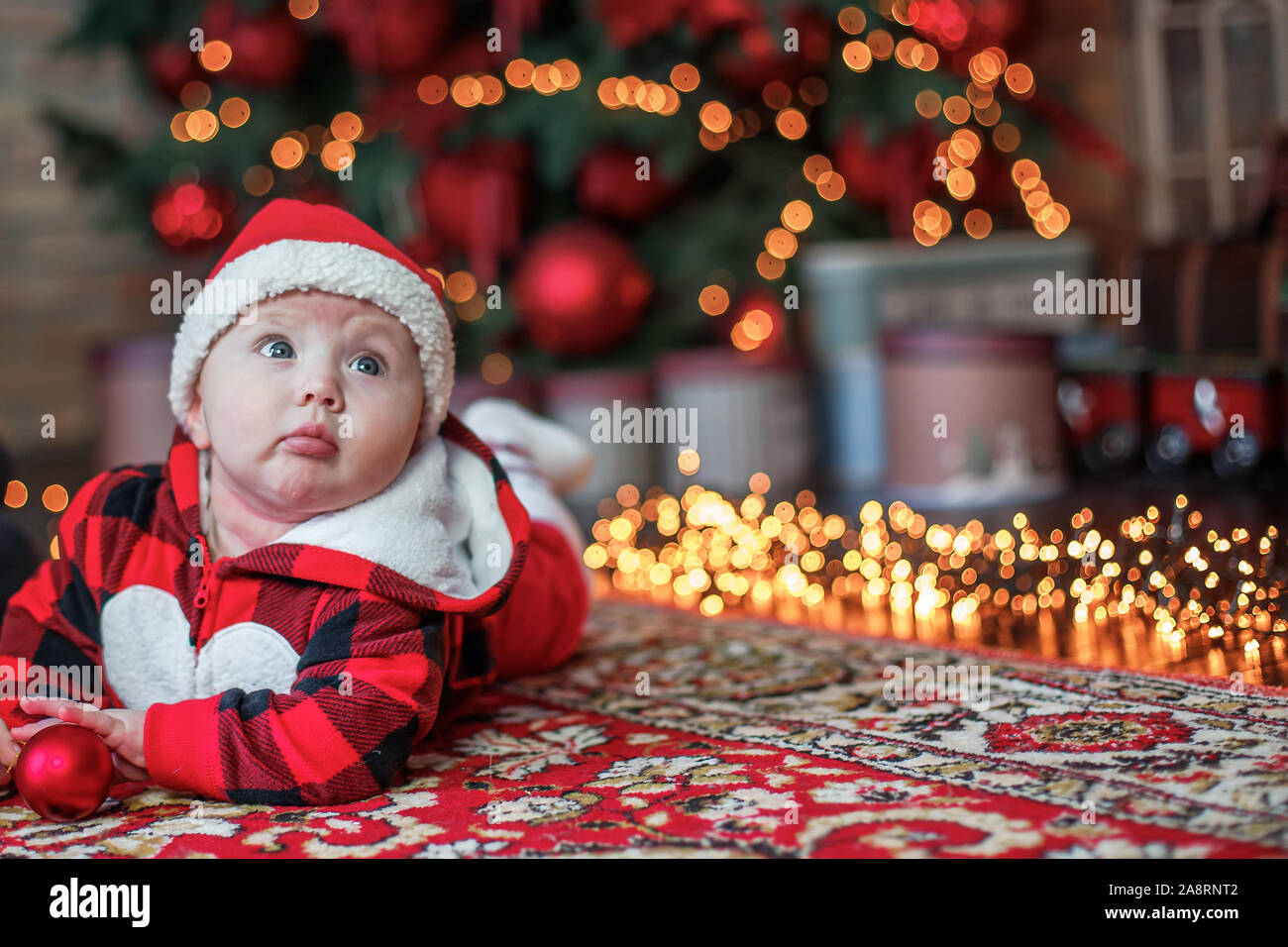 Little six month old baby dressed as Santa Claus. Background for christmas card. The child looks up at the place for inscription on background of lumi Stock Photo