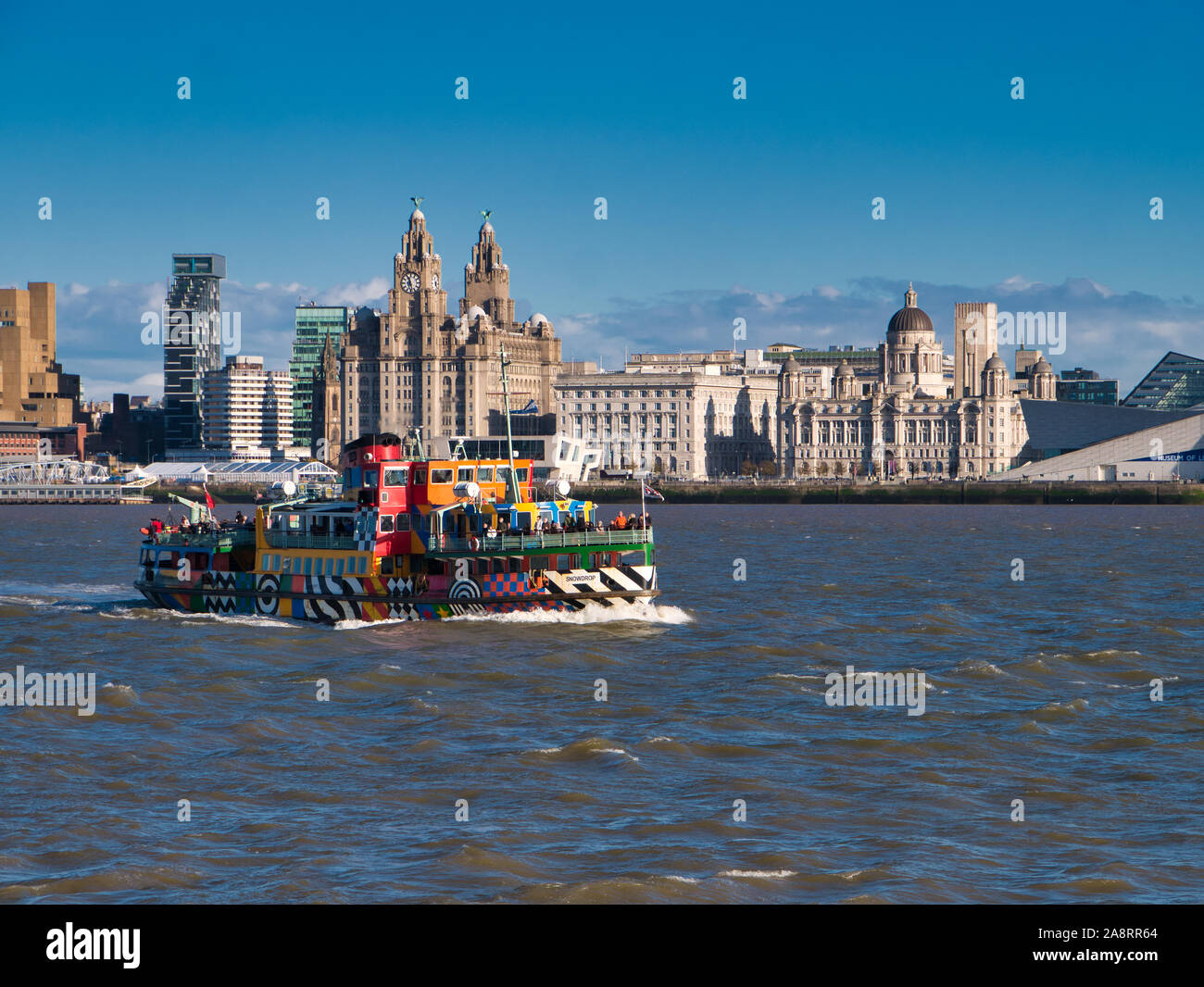 The River Mersey Ferry Snowdrop passes the Three Graces on the historic Liverpool waterfront on a sunny afternoon. Stock Photo