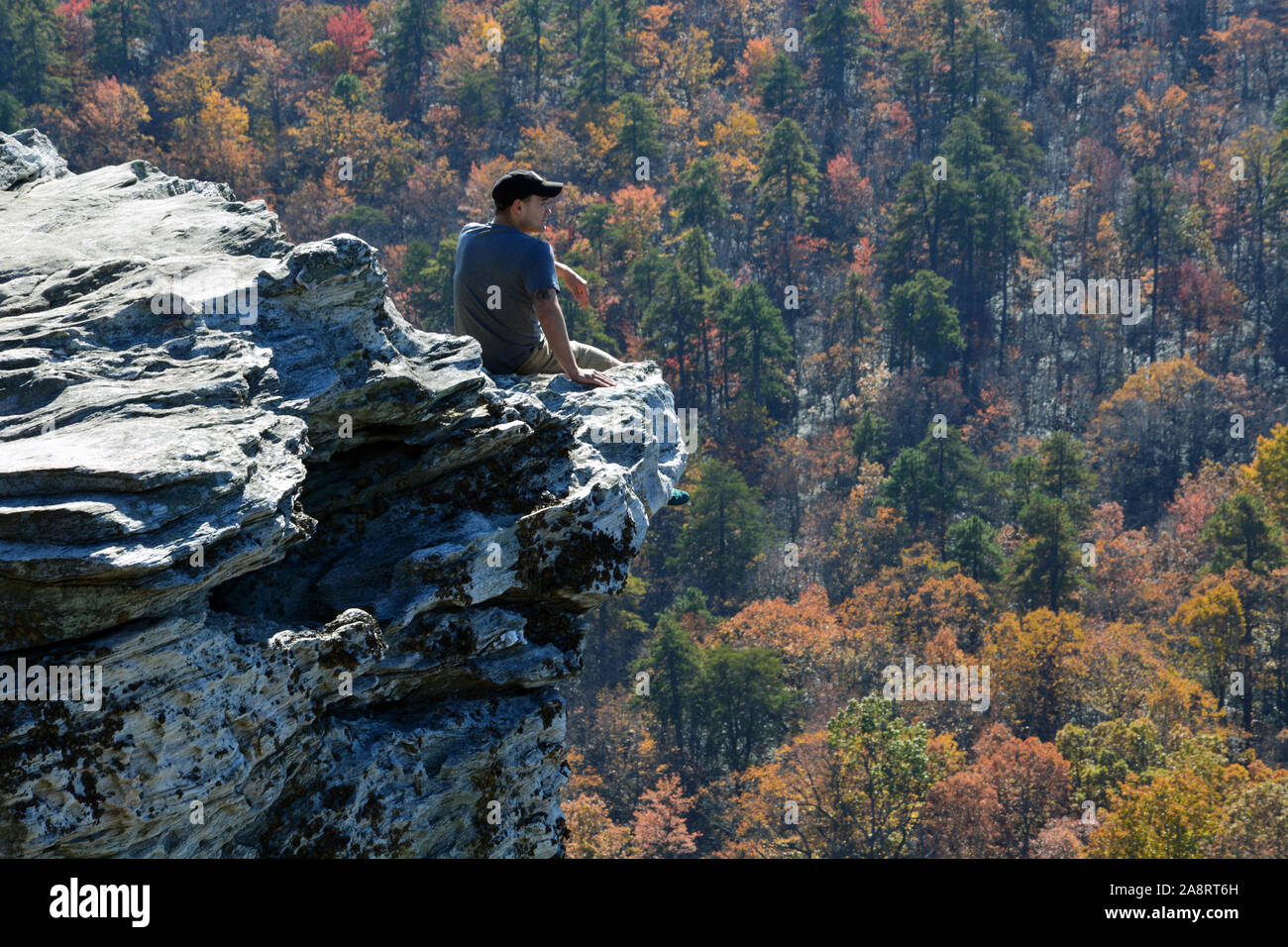 A young man sits perilously close to the edge, feet dangling over the cliff overlook, at Hanging Rock State Park outside of Danbury North Carolina. Stock Photo