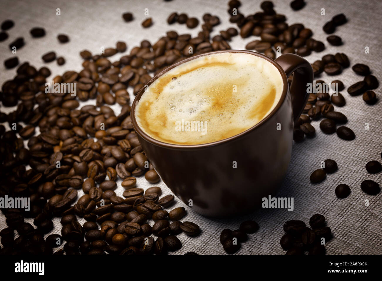 A cup of fresh coffee with foam on the table laid by burlap and scattered grains of coffee Stock Photo