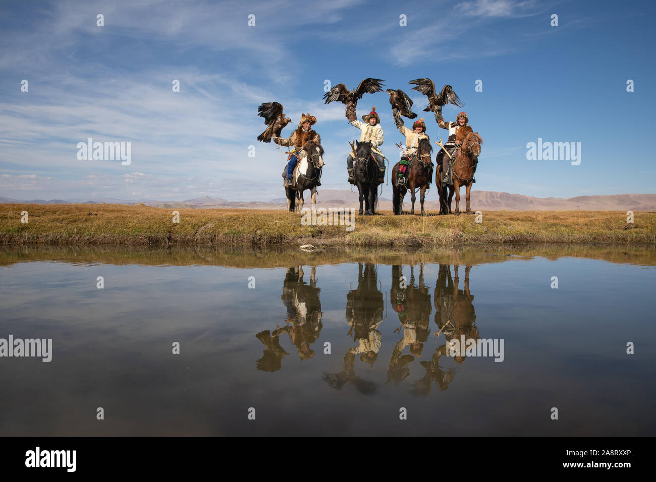 A group of traditional kazakh eagle hunters holding their golden eagles on horseback at the edge of the river with their reflections. Ulgii, Mongolia. Stock Photo